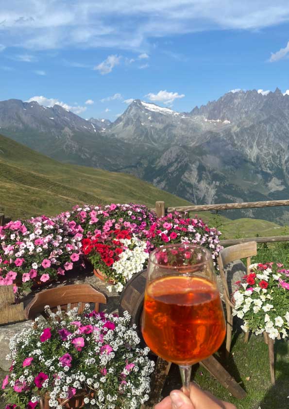 En primer plano hay un mocktail, de alquien que lo coge con la mano. En segundo plano hay muchas flores de color rosa y blancas, algunas son patunias. En un tercer plano hay unas montañas de los alpes italianos y se ve nieve de fondo.